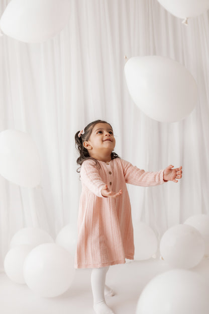 Child in a pink dress standing among white balloons against a white curtain.