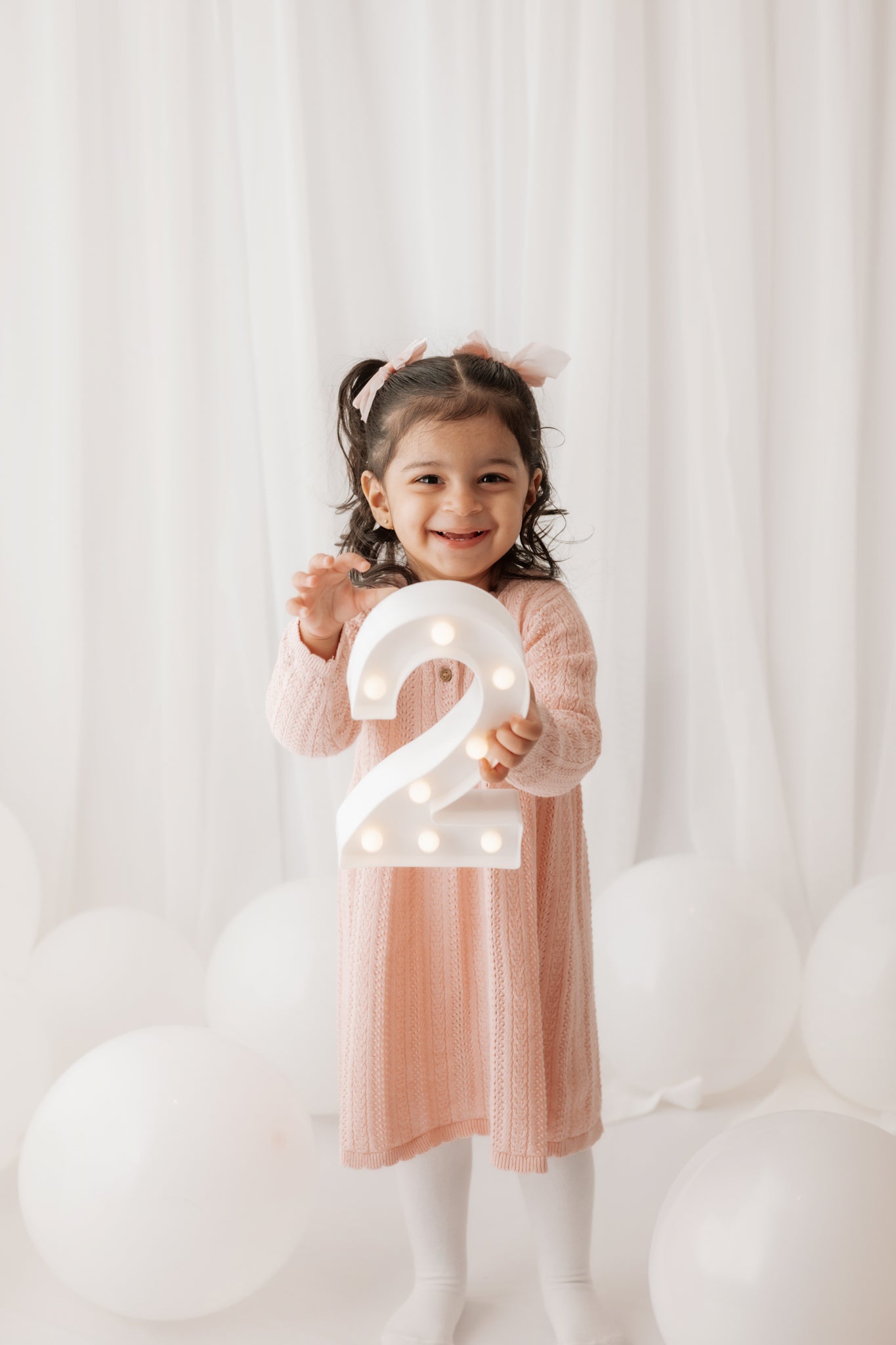 Child holding a number '2' light-up sign with white balloons in the background