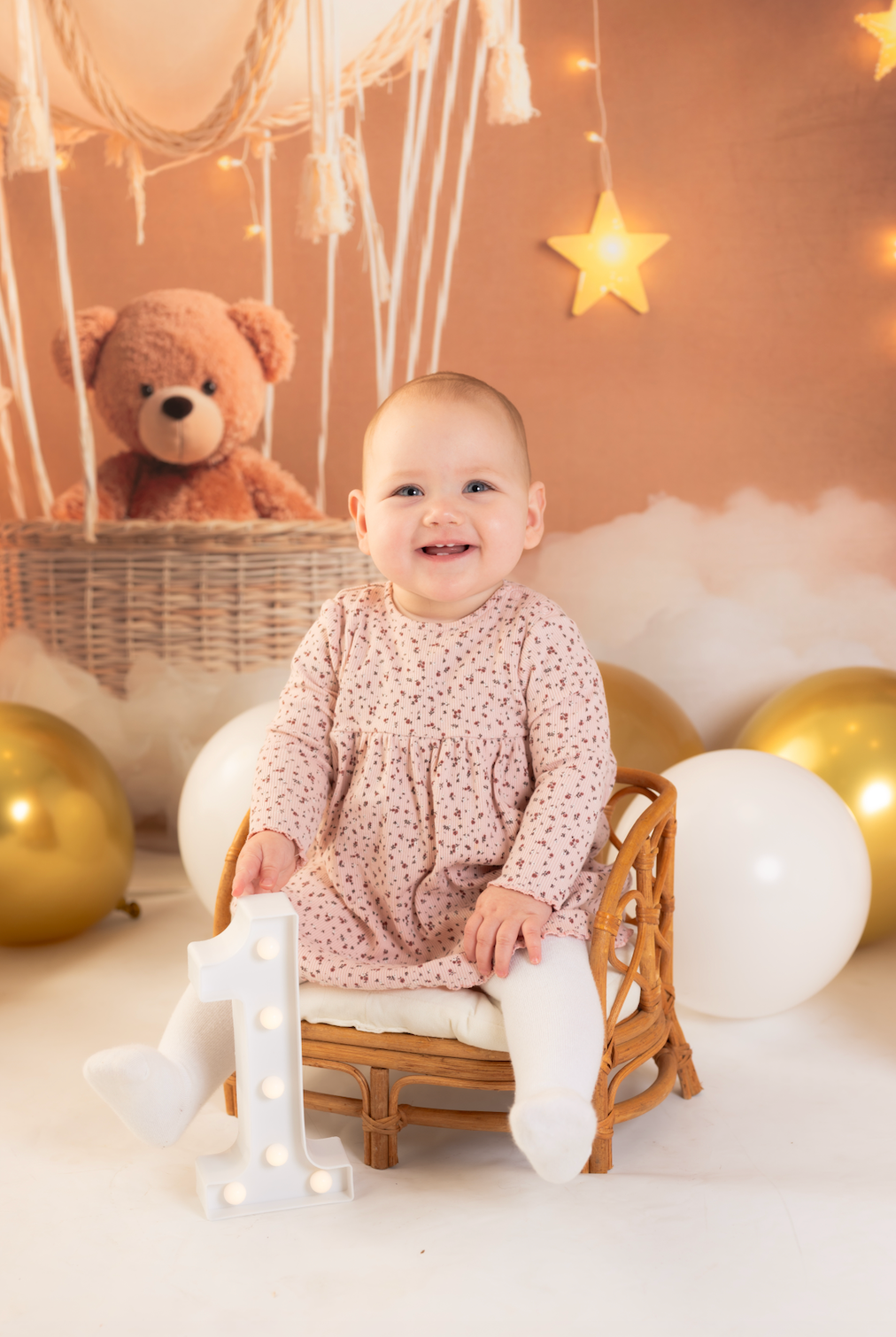 Child in a pink dress sitting on a small chair with balloons and a teddy bear in the background