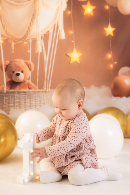Baby sitting on a white surface with balloons and a teddy bear in the background