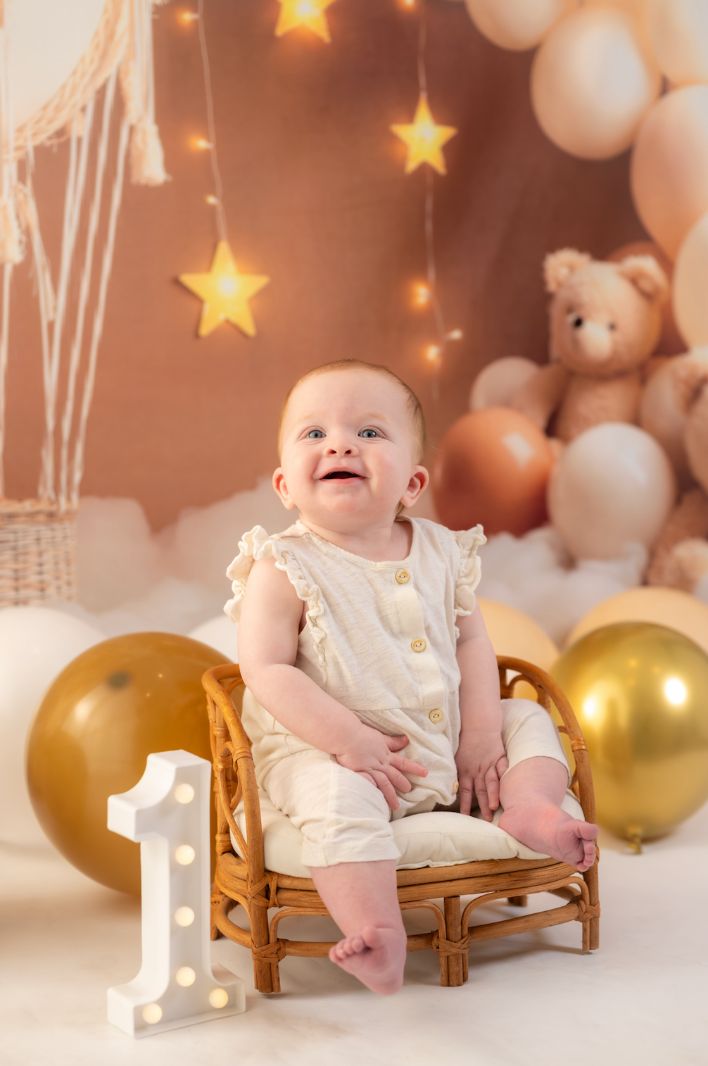 Baby sitting on a small chair with balloons and teddy bears in the background