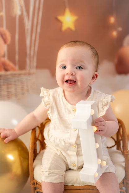Baby in a white outfit sitting on a chair with balloons and lights in the background