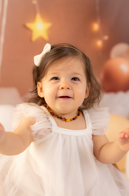 Child in a white dress with a decorative headband and necklace, surrounded by warm lighting and blurred balloons.
