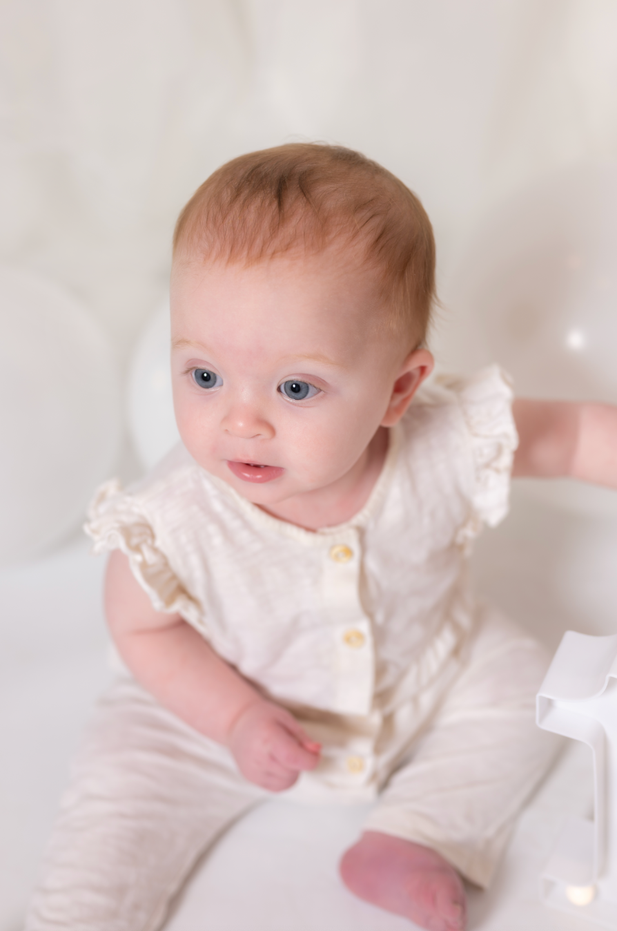 Baby in a white outfit with ruffled sleeves sitting on a white surface.