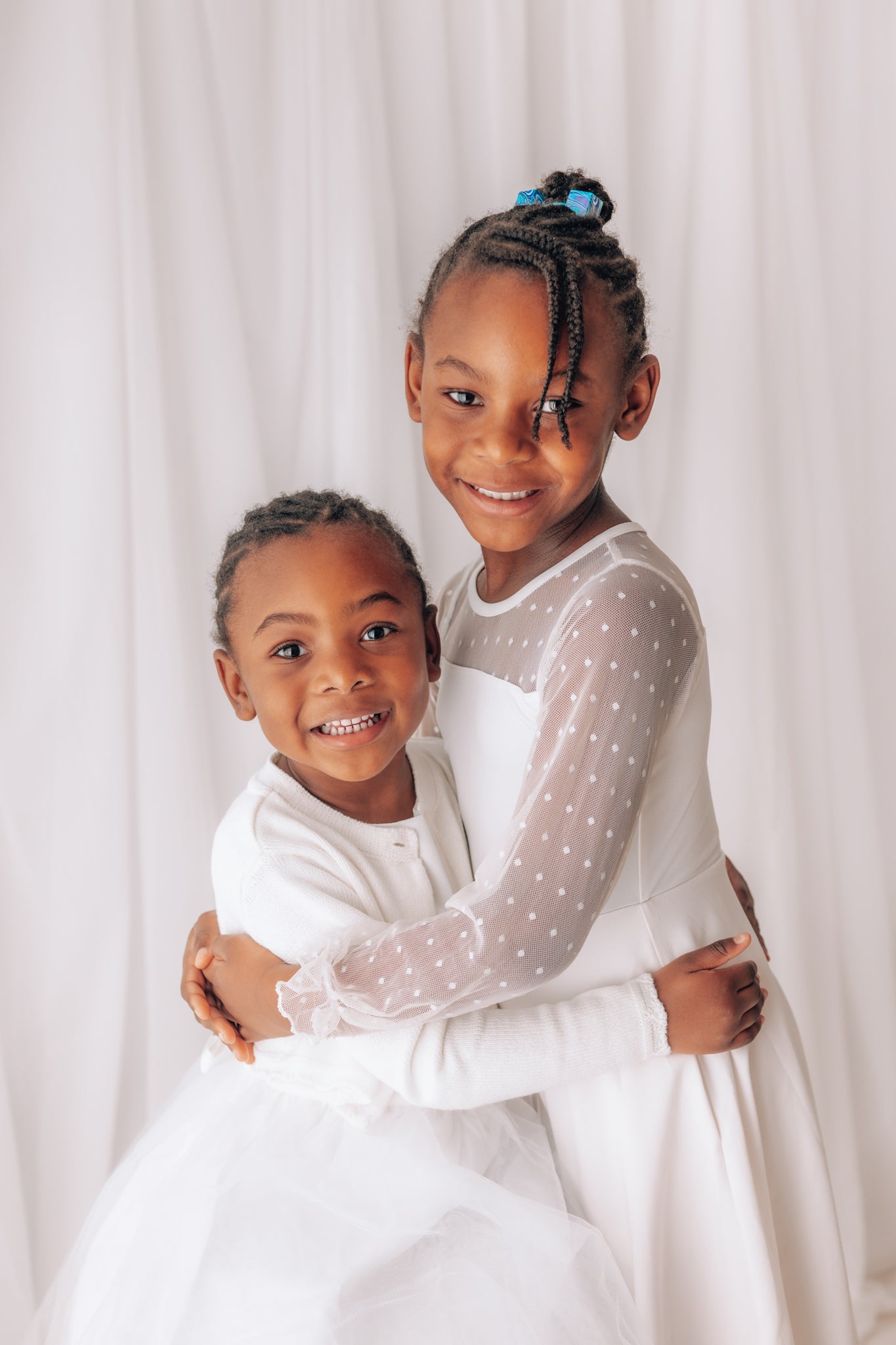 Two children in white dresses hugging against a white background