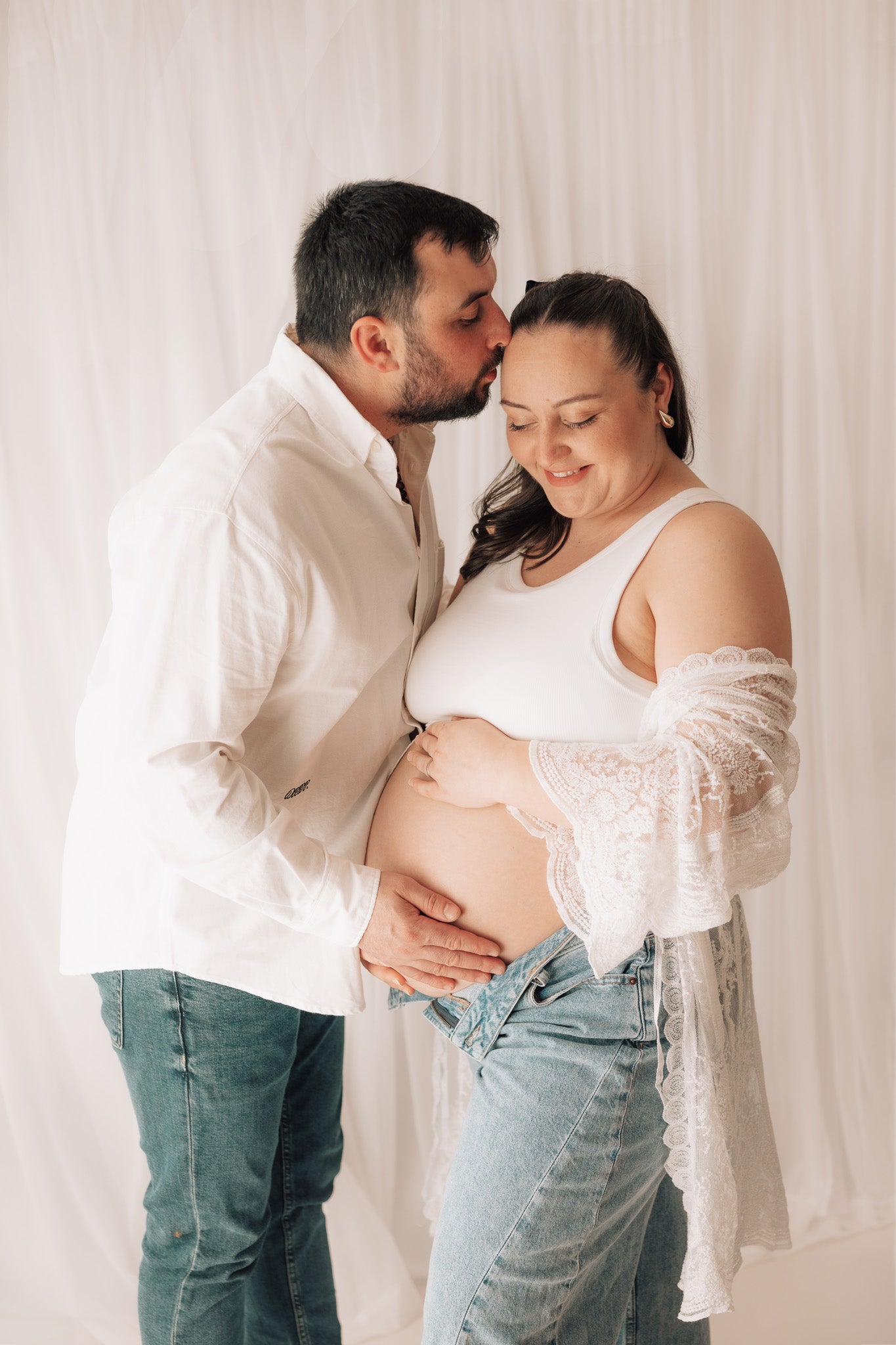 Pregnant woman with a man in a white shirt and jeans, standing close together against a white background.