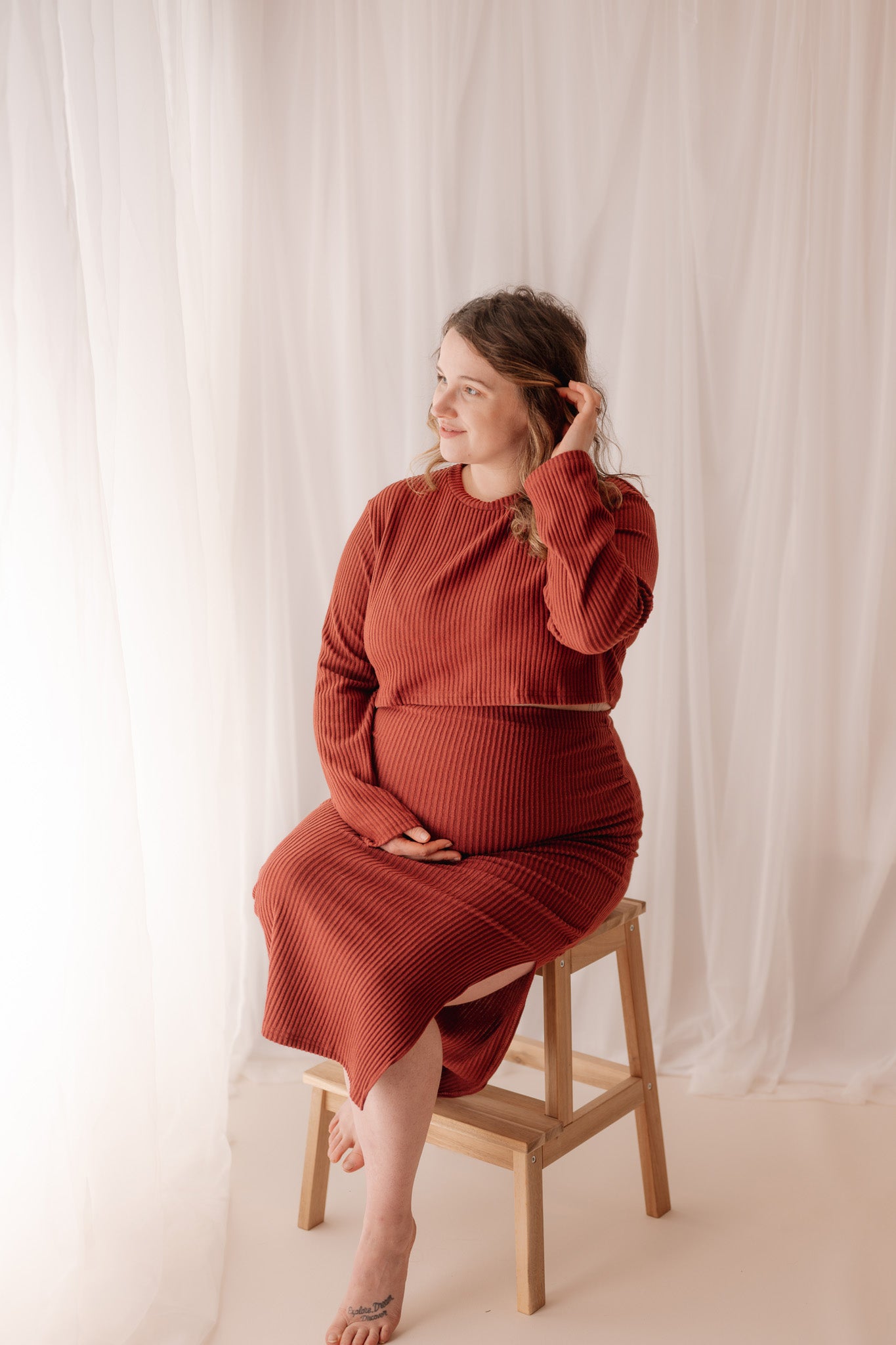 Woman in a red outfit sitting on a wooden stool against a white curtain during a Surrey maternity studio photoshoot