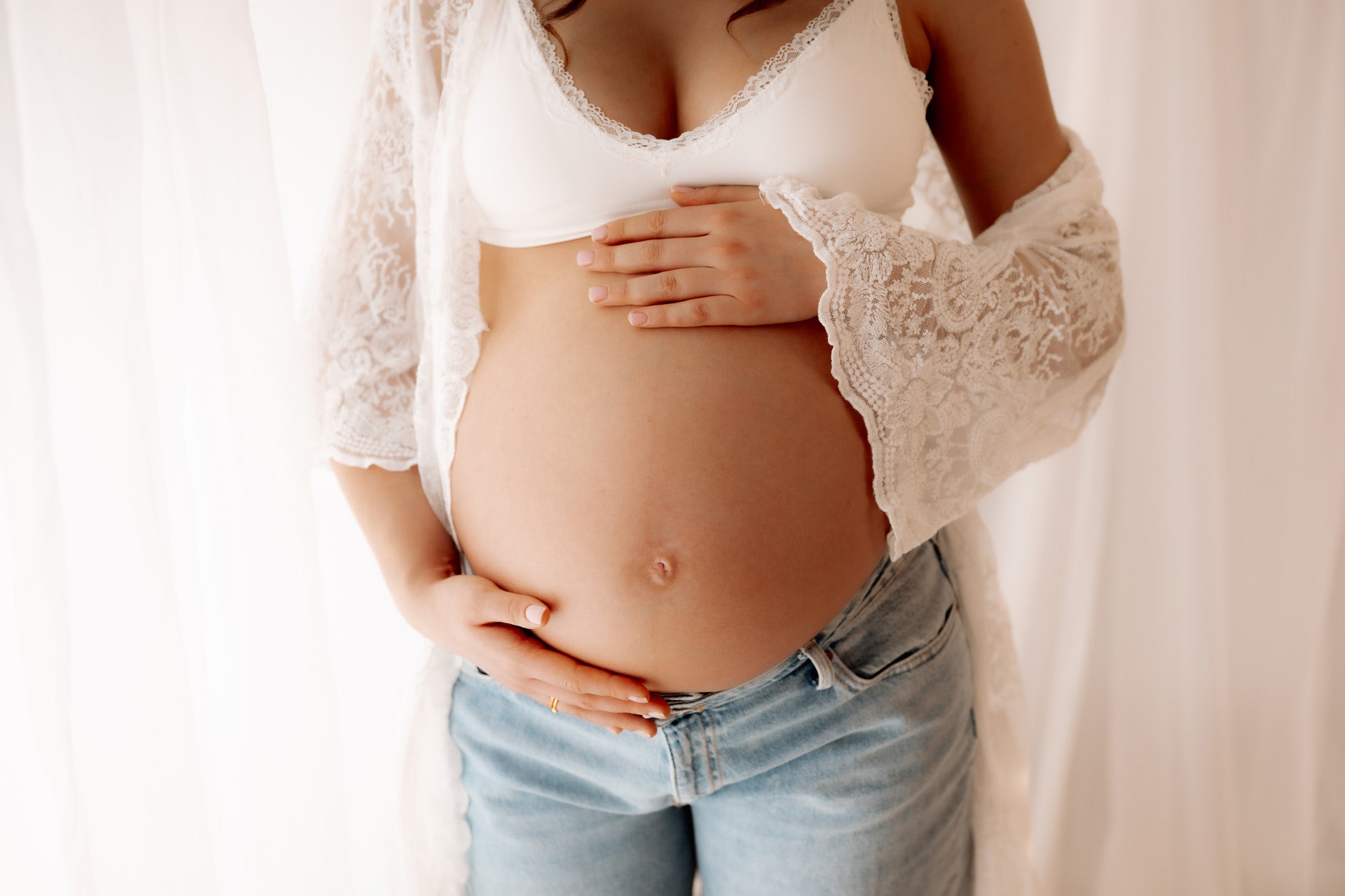 Pregnant person wearing a white lace top and blue jeans against a plain background in Surrey photography studio