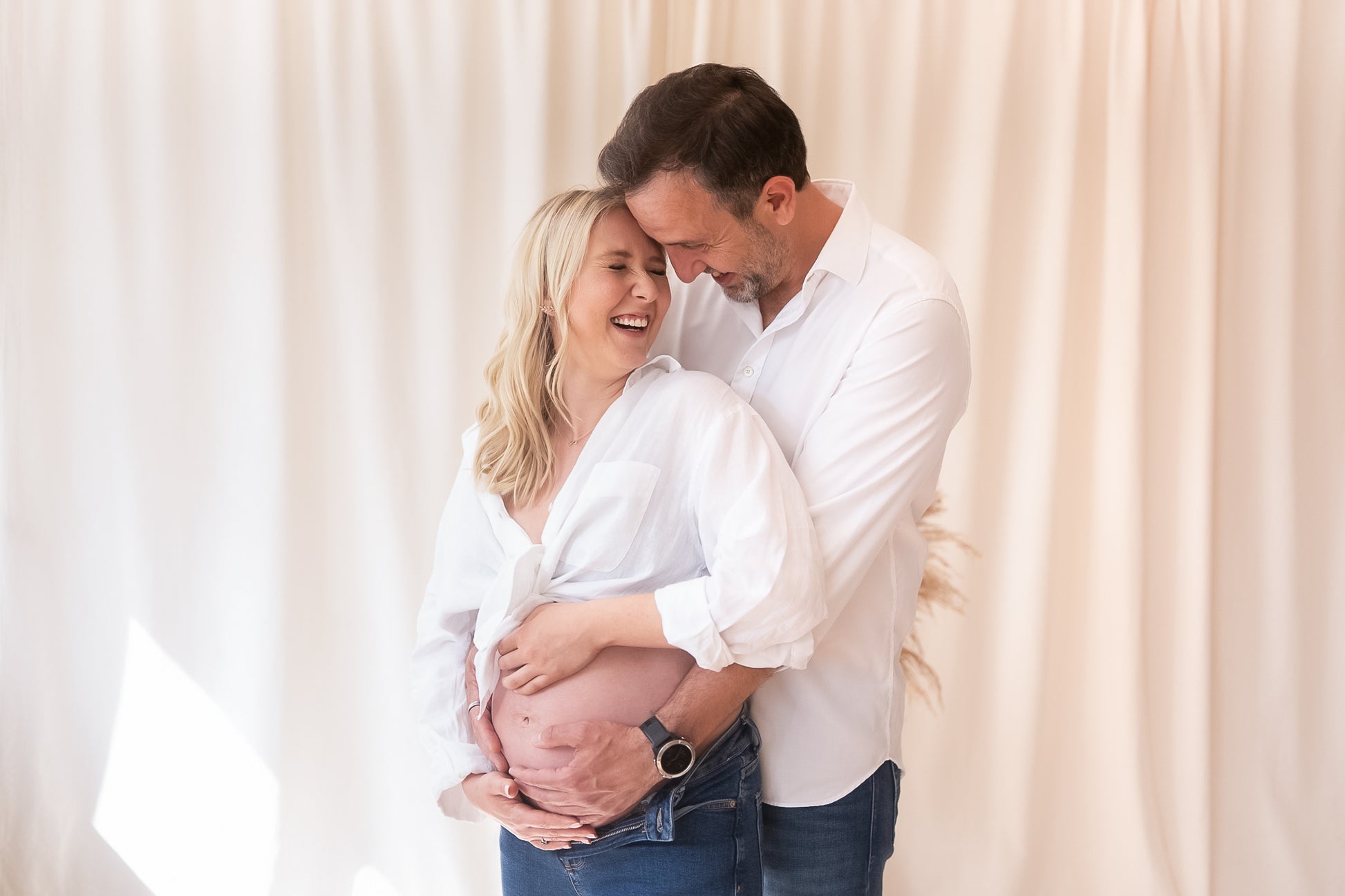 Pregnant woman being embraced by a man against a white curtain background