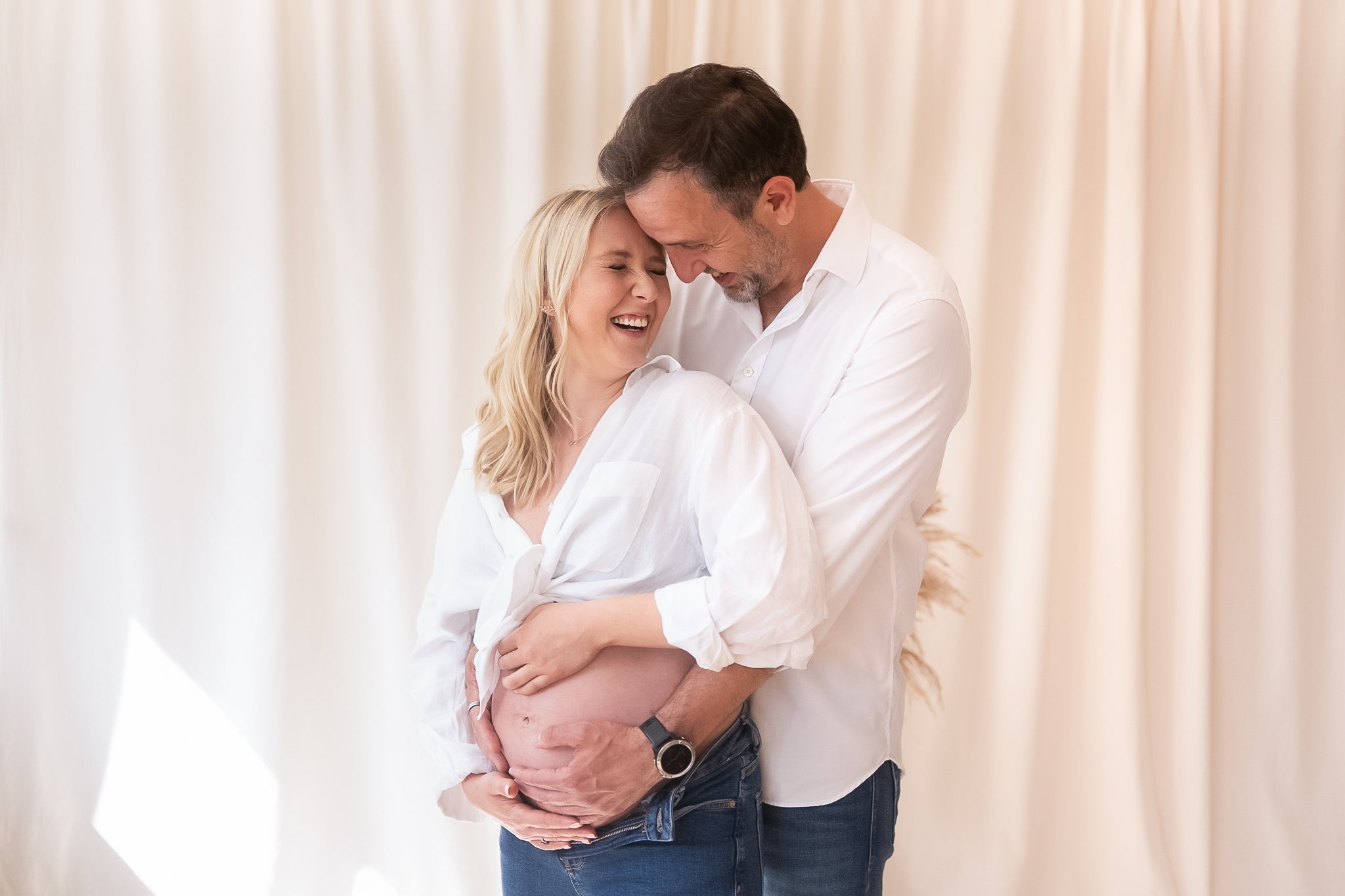 Pregnant woman being embraced by a man against a white curtain background