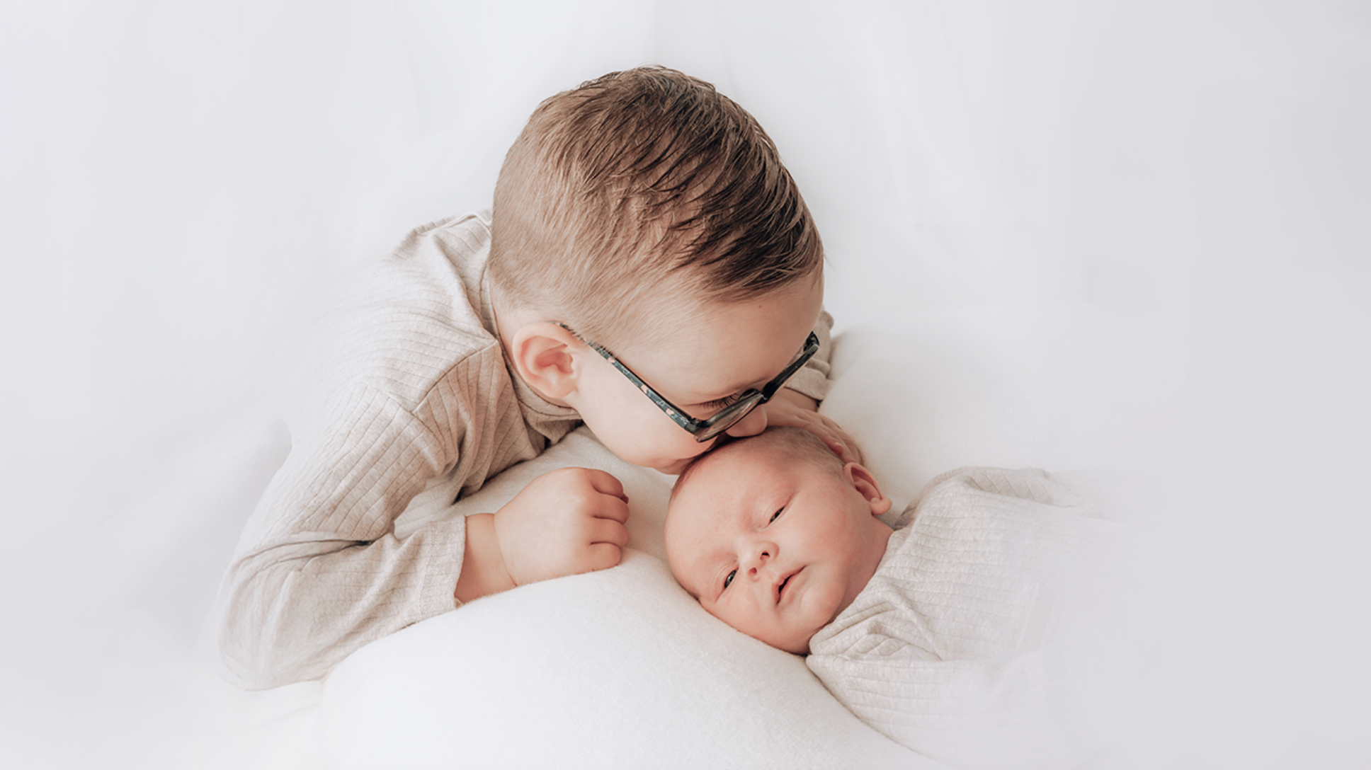 Two children lying on a white blanket, one wearing glasses during a studio photoshoot for families and babies