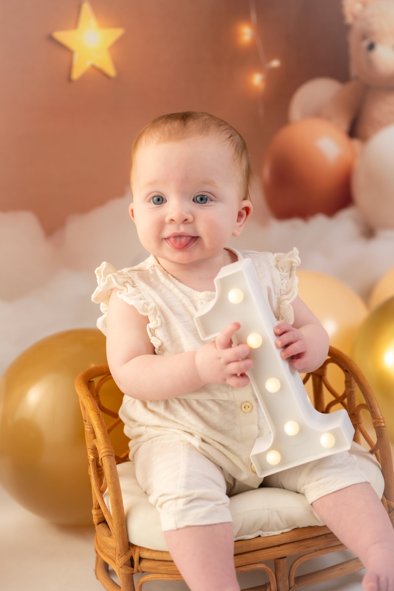 Baby sitting on a chair holding a light-up letter 'A' with balloons and a teddy bear in the background.